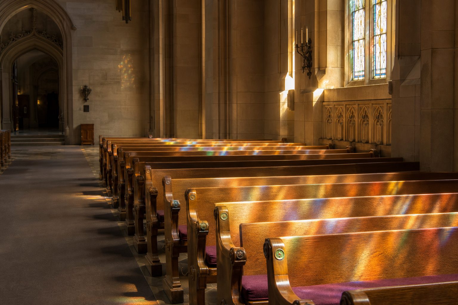 Rows of empty wooden pews with plum cushions in a stone sanctuary, stained glass color falling across the seats.