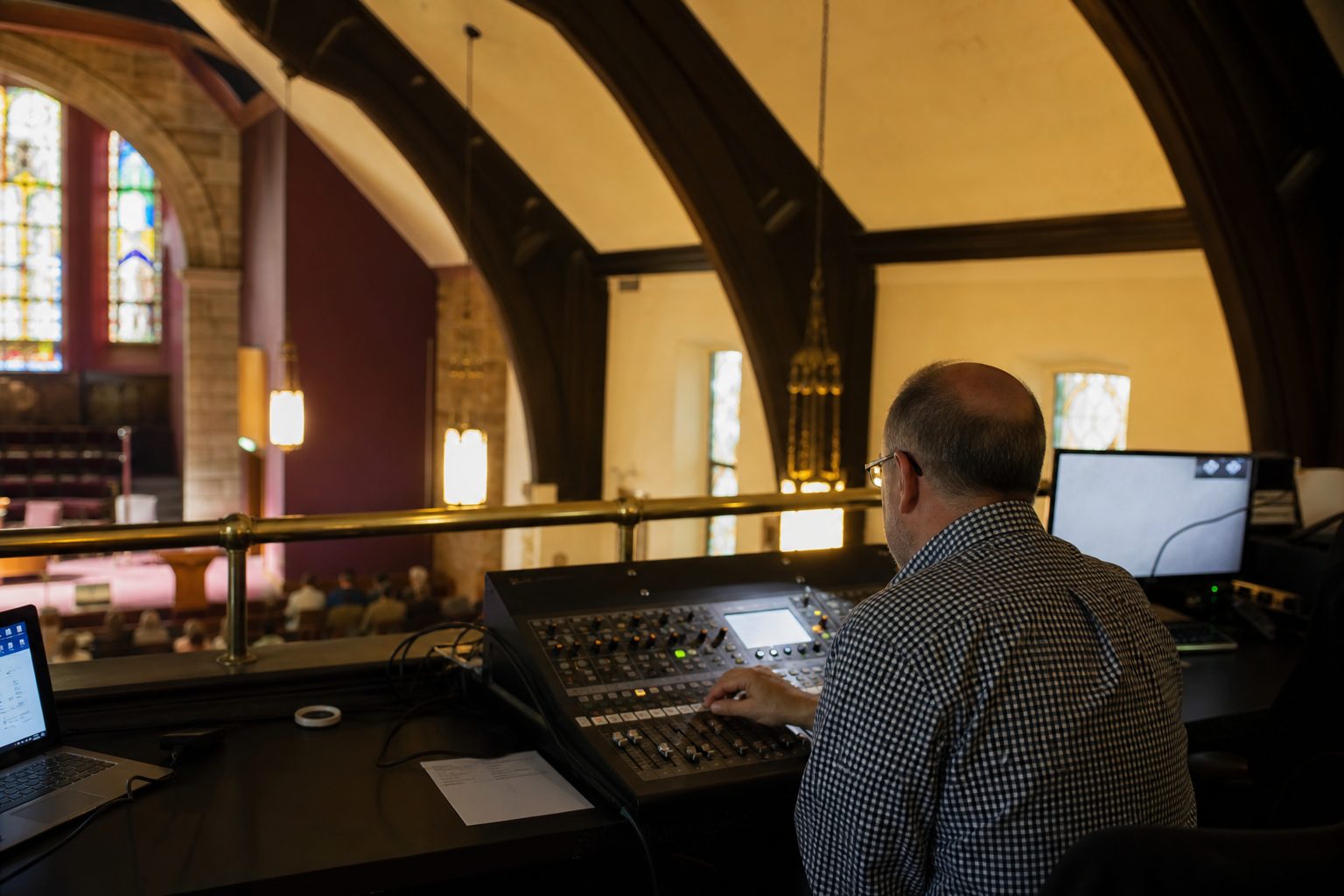 Volunteer at a small audio mixing desk in a church tech booth at the back of a stone sanctuary.