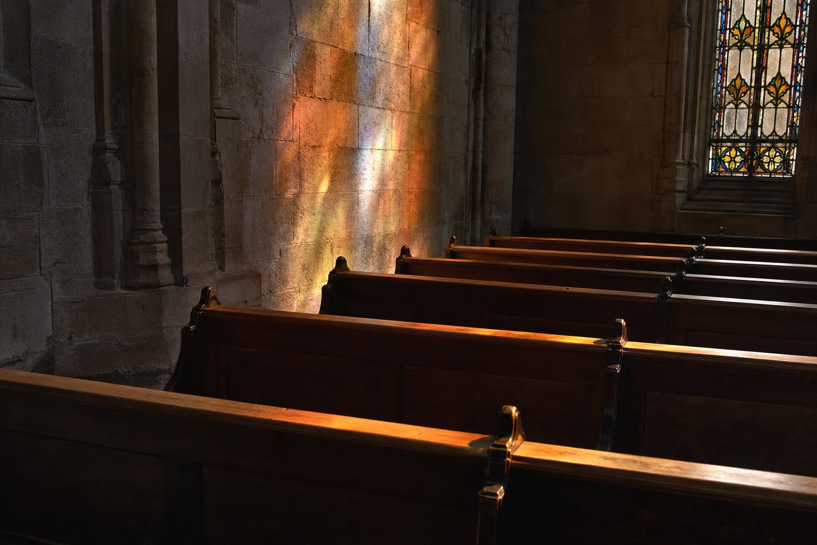Empty wooden church pews with warm stained glass light falling across them in a quiet stone sanctuary